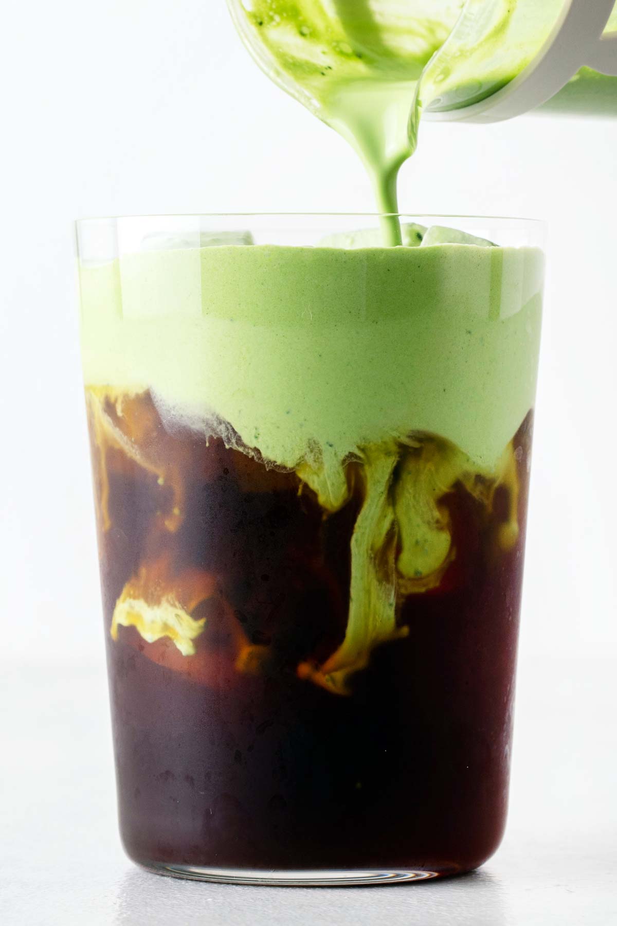 Matcha koudschuim wordt over een ijskoffie in een groot glas gegoten.